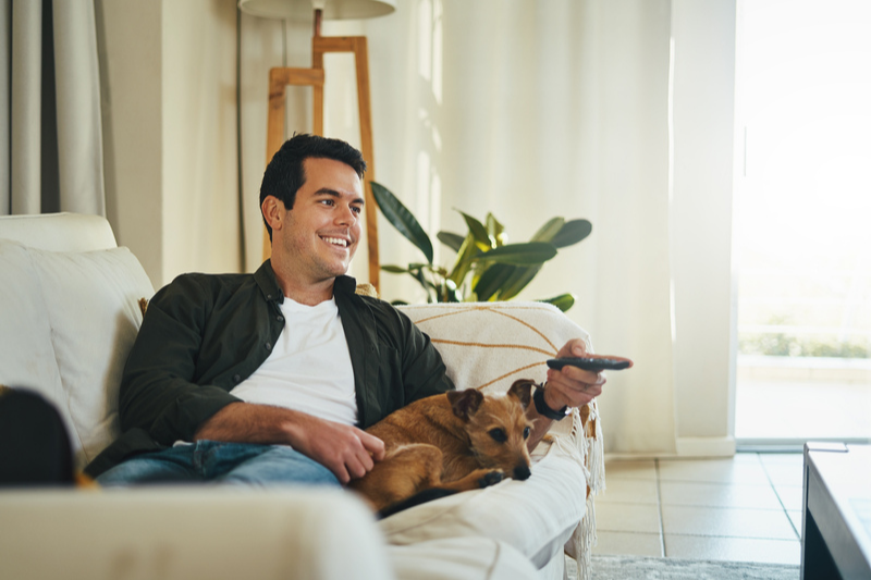 Smiling man relaxing on couch with his dog holding remote control in hand watching tv in living room.